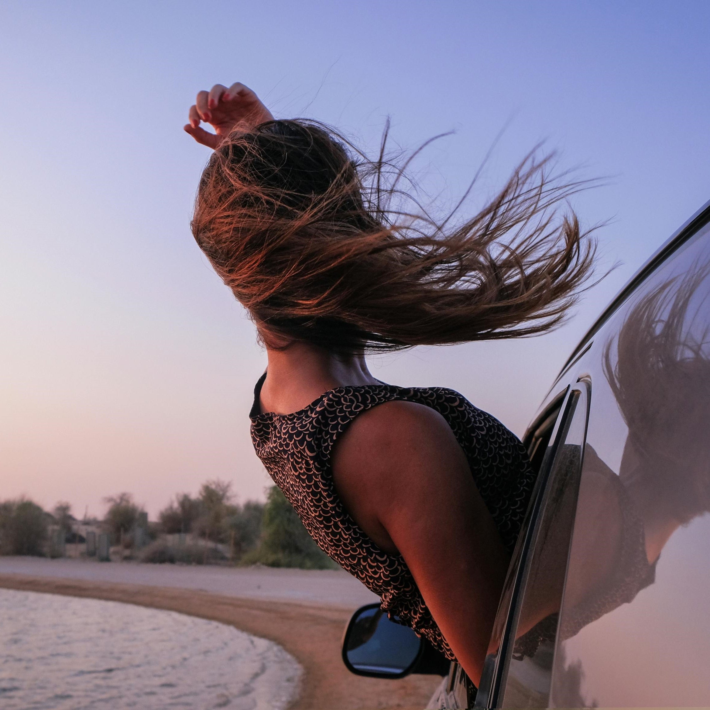 Carefree moment of a woman leaning out of a car window, her hair flowing in the wind against a serene backdrop of a beach and pastel sunset, capturing a sense of freedom and adventure.
