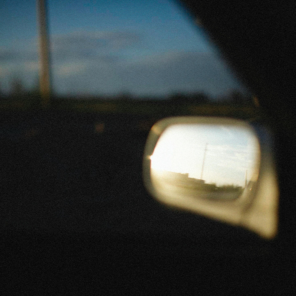 Artistic shot of a car's side mirror reflecting a sunlit scene against a blurred, moody background, symbolizing fleeting moments and the beauty of travel.