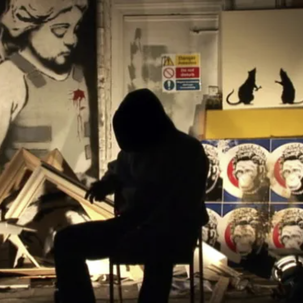 ilhouette of a hooded figure sitting on a chair in front of urban street art, featuring a distressed child image, a warning sign, and a Banksy-style artwork with two cats and posters of a monkey figure.