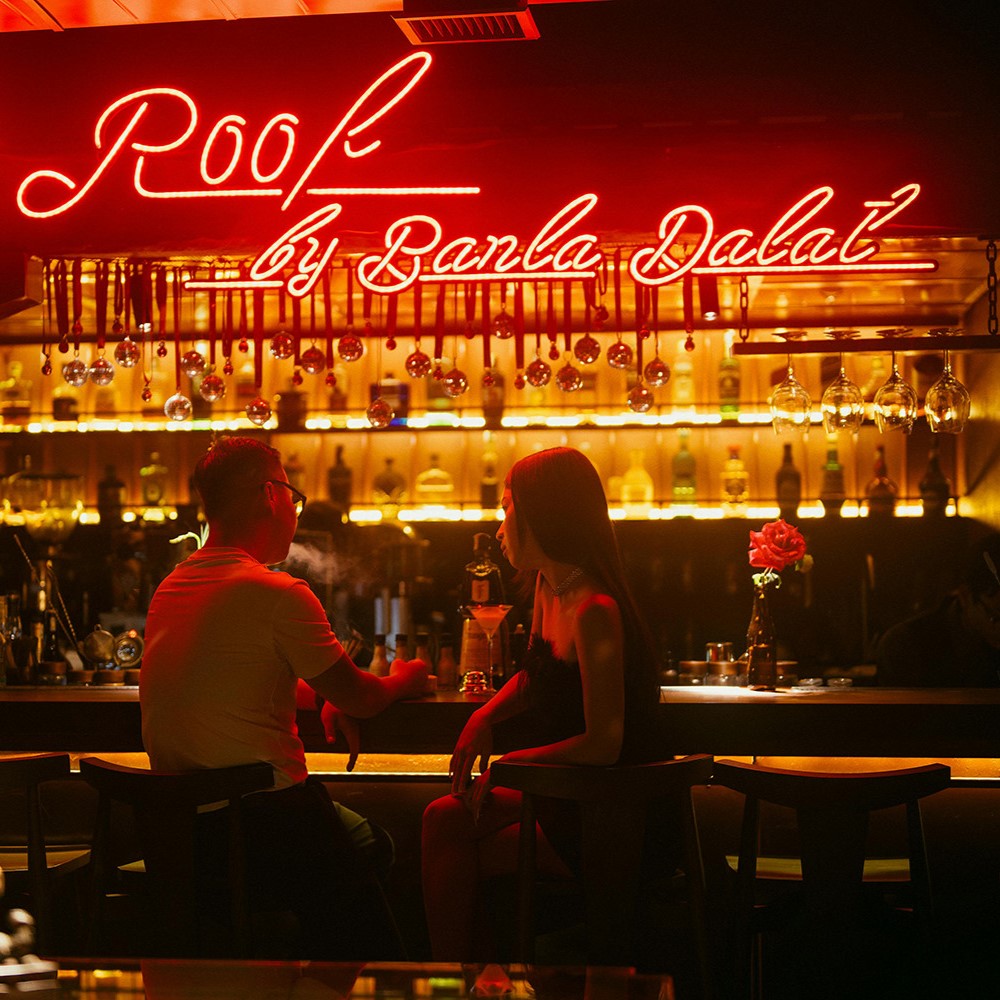 Moody bar scene featuring a man and woman sharing an intimate conversation under warm neon lighting, with a backdrop of glowing bottles and hanging glassware, creating a romantic and sophisticated ambiance.