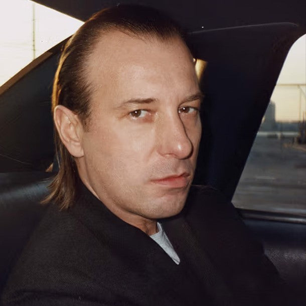 Close-up portrait of a man with slicked-back hair, sitting in a car and staring intently at the camera with a serious expression.