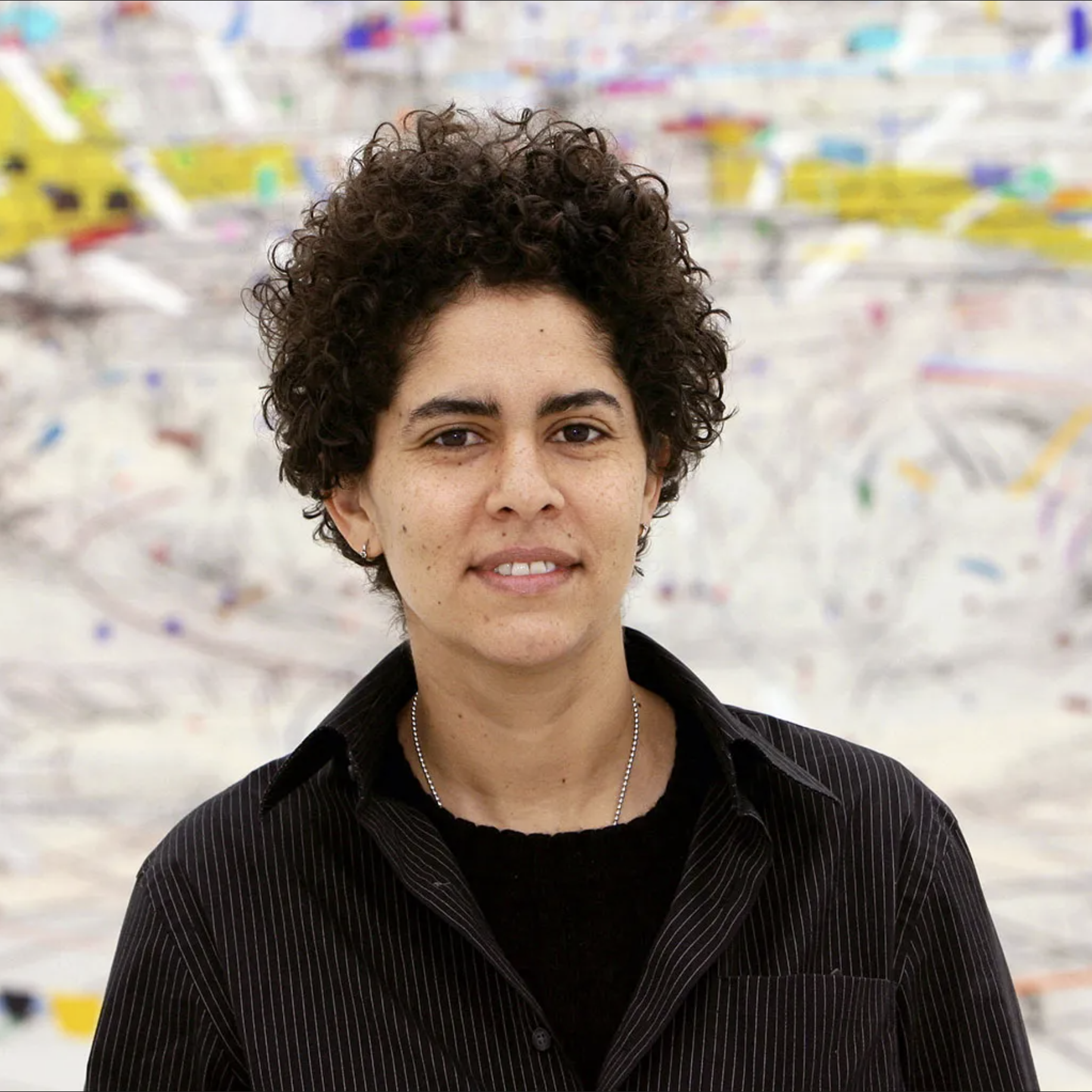 Portrait of artist Julie Mehretu standing in front of one of her colorful abstract artworks, with short curly hair and a black striped shirt.