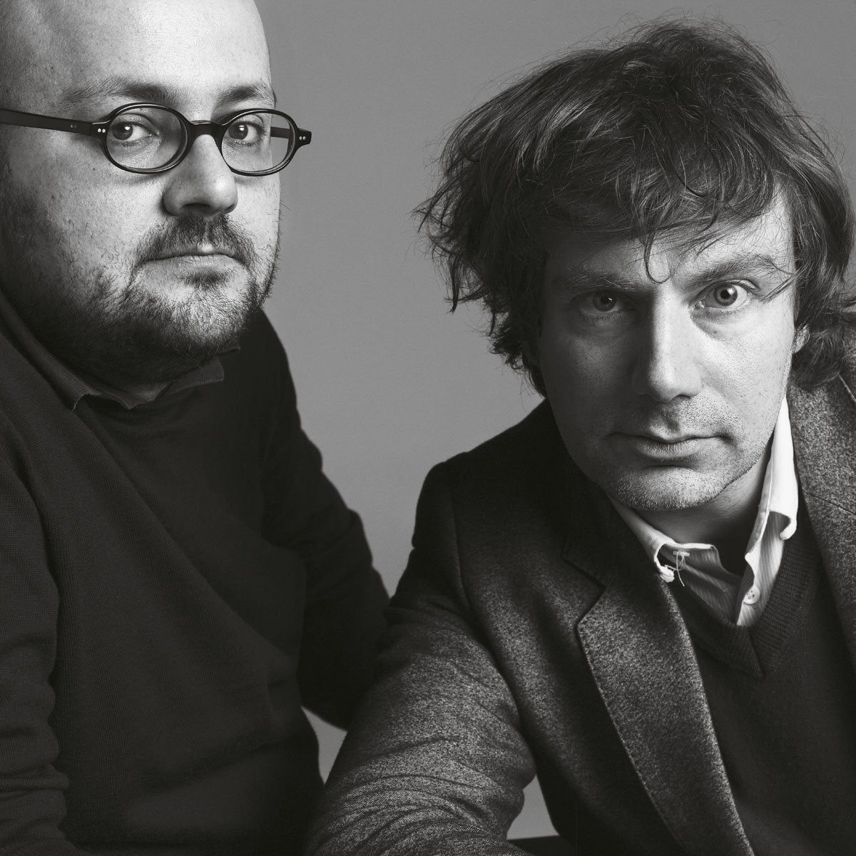 Black-and-white portrait of two men, one with round glasses and a dark sweater, and the other with messy hair and a blazer, both staring directly at the camera.