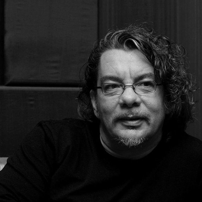 Black-and-white portrait of a man with curly hair, glasses, and a beard, sitting with a thoughtful expression against a textured dark background.