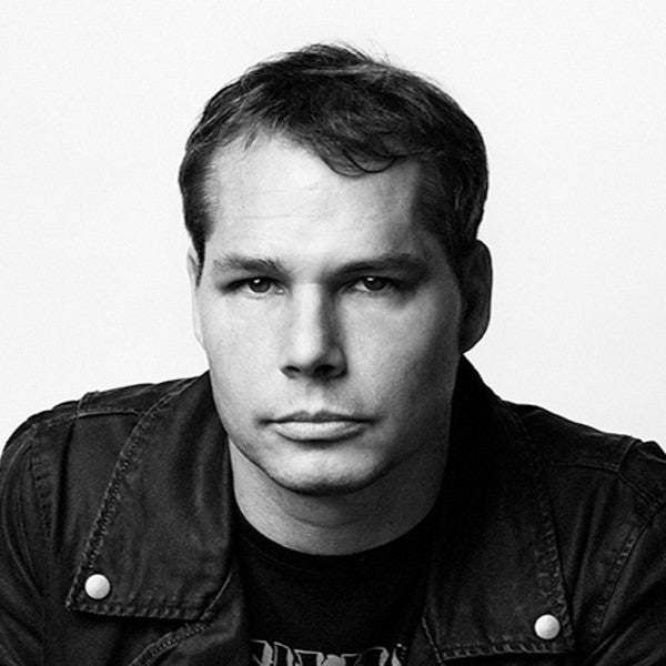 Portrait of artist Shepard Fairey, known for his iconic street art and political posters, standing in front of a mural with bold, graphic design elements in red and black.