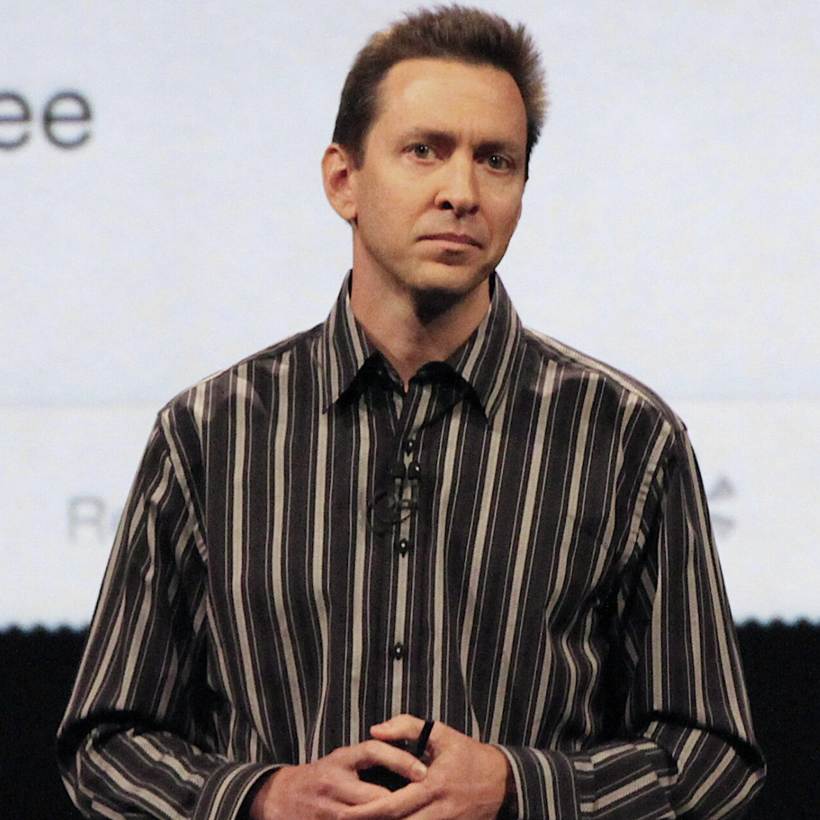 Portrait of Scott Forstall, former Apple executive and key architect of iOS, smiling and wearing a suit in front of a tech-themed background.