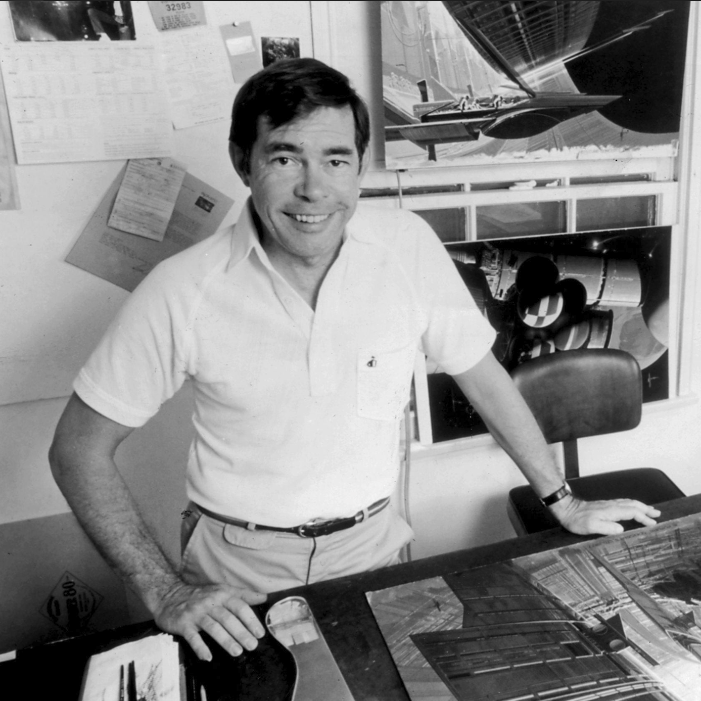 Black-and-white photo of conceptual artist Syd Mead standing at a desk with sketches and futuristic designs in the background, smiling and dressed in a white shirt and khaki pants.