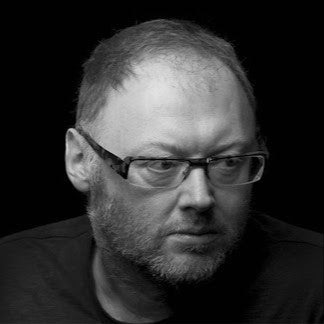 Black-and-white portrait of a man with glasses and a beard, looking thoughtfully to the side against a dark background.