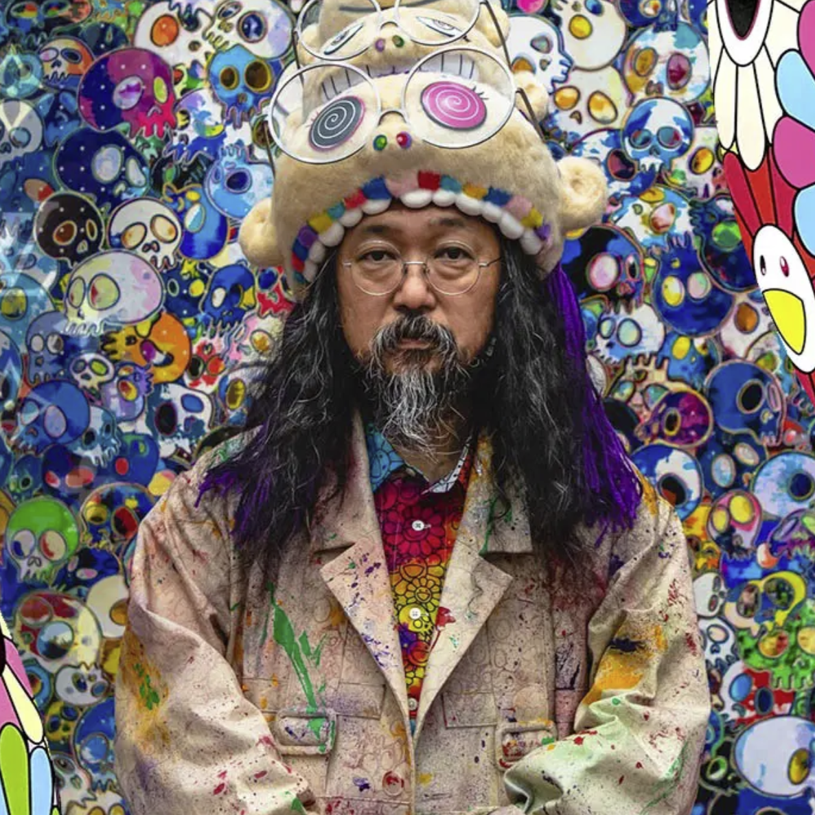 Portrait of Takashi Murakami standing in front of a vibrant, colorful backdrop of skull and flower artwork, wearing a playful, oversized hat with large glasses and a colorful painted coat.